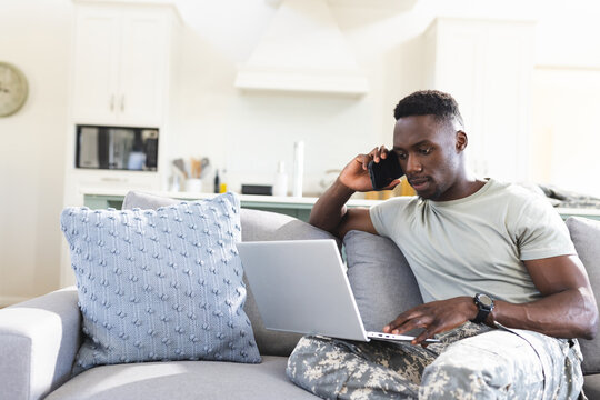 African american male soldier on couch at home using laptop and talking on smartphone, copy space