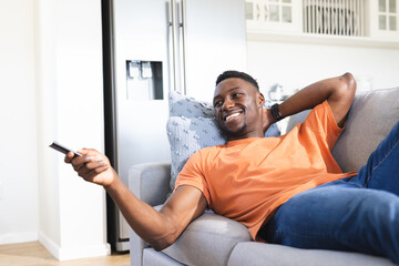 Happy african american man relaxing on sofa watching tv, using remote control