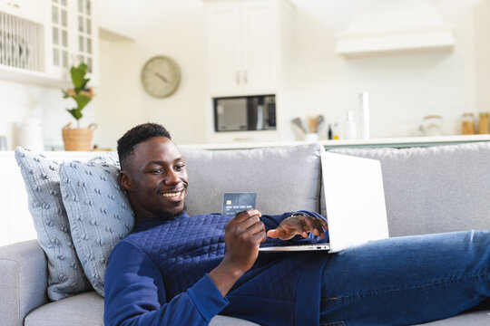 Happy african american man relaxing on sofa using laptop and credit card online, copy space - Powered by Adobe