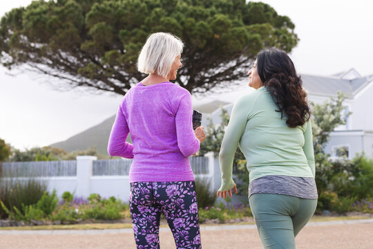 Rear view of two happy diverse senior female friends walking for exercise in country road, talking