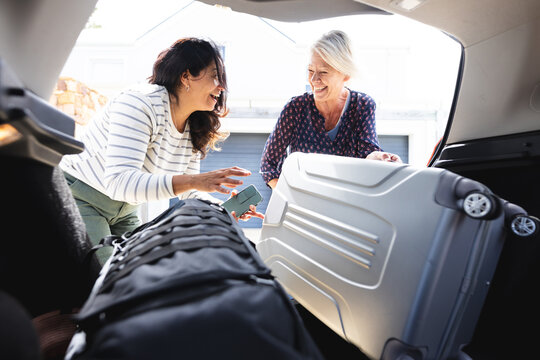 Two happy diverse senior female friends going on trip packing car boot with luggage, copy space