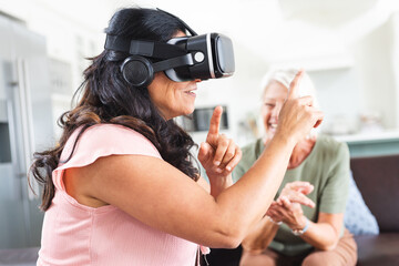 Two happy diverse senior female friends laughing and using vr headset at home