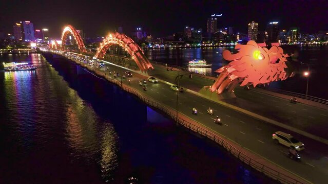 Dragon Bridge in Da Nang, Vietnam, beautifully illuminated at night. The scene captures various vehicles crossing the bridge, while boats navigate the river below.