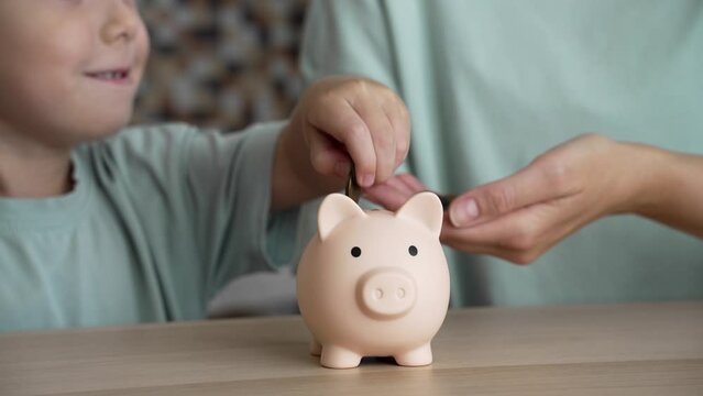 A mother teaches her son about saving money by having him put coins into a piggy bank. Financial literacy education from an early age. A child and his mother save money together.