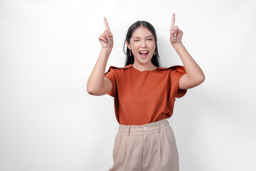 Excited young Asian woman in a brown shirt and taupe pants pointing to the copy space above her,...