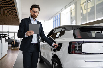 Salesman in suit sells electric car in dealership using tablet. Promoting sustainable...