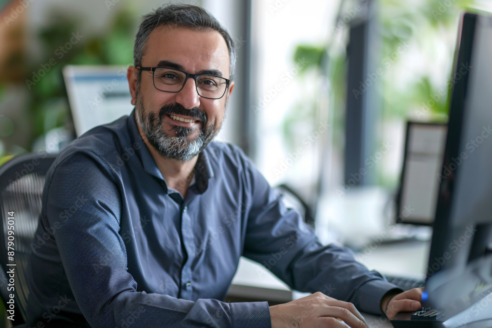Poster portrait of a smiling businessman sitting at a desk with a computer and looking at the camera, with 