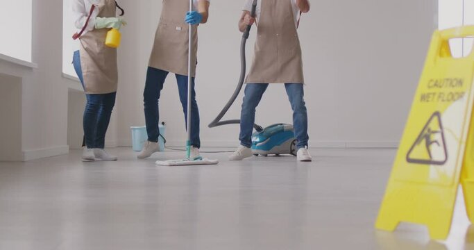 Cleaning. Cheerful cleaning crew playfully dances while working next to bright yellow 'Caution Wet Floor' sign. Group of cleaners in aprons having fun in empty room with mops and vacuum cleaner.