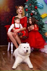 Family photo portrait. Mom and her two children and a white dog in red clothes celebrate the Chistmas, new year. Happy young mother and her daughter, son and a dog in a cozy dark living room on