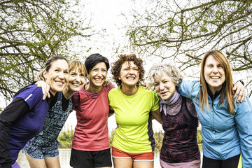 Group of smiling women of various ages in athletic wear enjoying outdoor exercise together. They are embracing each other in a park setting. Concept of friendship, fitness, and healthy lifestyle.