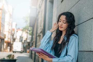 A female student with a backpack smiles and holds a notebook, a folder in her hands doing homework outside of her. The concept of education, study, exams.