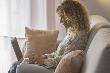 Blonde woman with curly hair using laptop sitting on sofa at home. 45 year old serious middle aged...