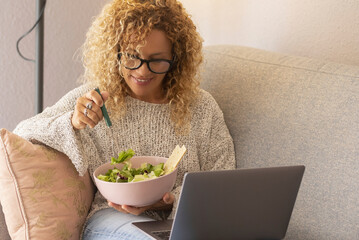 Blonde woman with curly hair using laptop sitting on sofa at home. Middle-aged smiling female of 45...