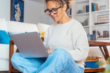 Modern middle age woman writing on computer sitting cross legs on the floor at home in relaxation...