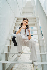 One young caucasian woman student sitting on stairs near university listening to music and drinking coffee	