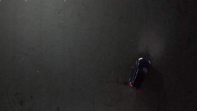 An aerial top down view of a blue SUV driving recklessly and doing doughnuts in an empty parking lot at night. The drone camera is tilted straight down, directly over the vehicle.