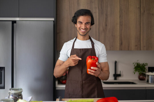 Smiling man in apron pointing at a red bell pepper in a modern kitchen.