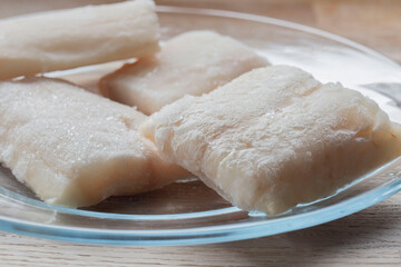 Frozen haddock fillets defrosting on a glass plate. On a wooden chopping board background