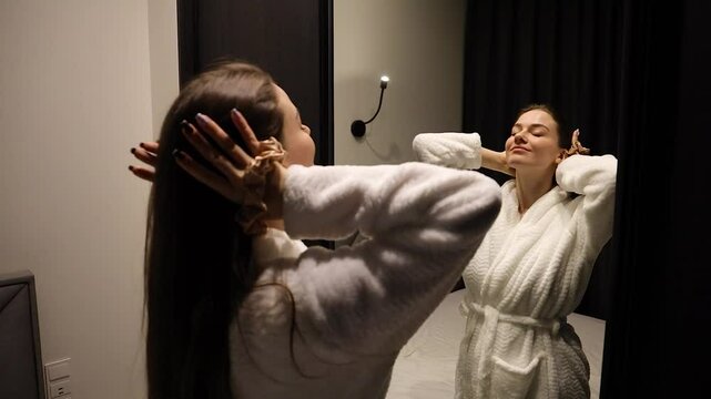 Pretty young attractive woman tying her hair with a scrunchie while standing in front of a mirror wearing a white robe