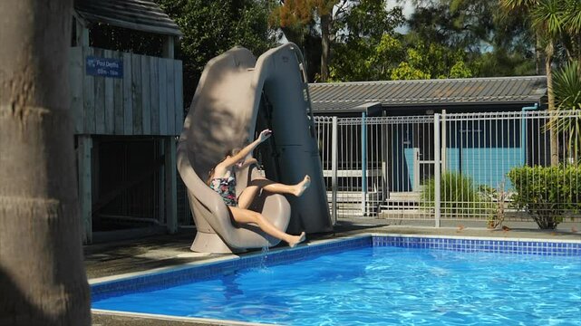Preteen girl playfully slides down a water slide and splashes in slow motion into a pool of water