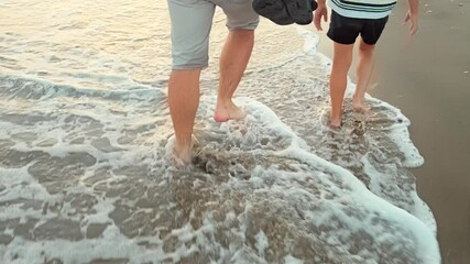 Father and son walking together along the seashore, the feet are washed by the sea waves close-up. Family walk together. Side view. Parents and children relationships. Love concept. Happy time. 4K