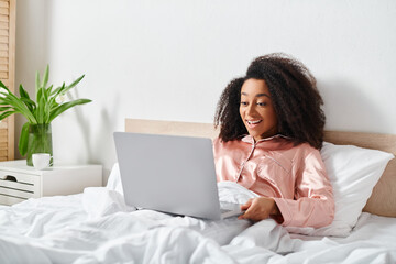 African American woman in pajamas sitting on bed, using laptop in bedroom in the morning.