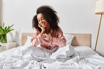 A curly African American woman in pajamas sits calmly on a bed with white sheets in a serene...