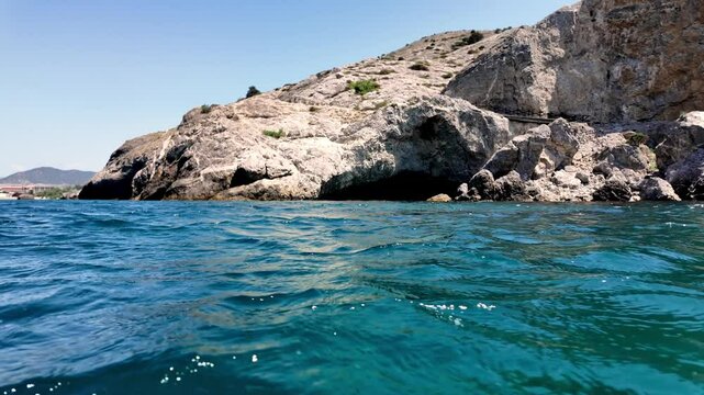 A view of the coastline in Sudak, Crimea, Russia. The camera is facing the sea, with rocky cliffs and a cave in the background.