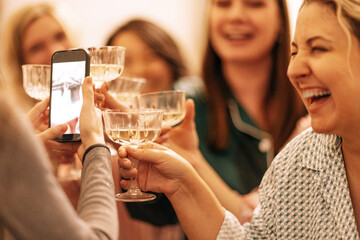A close-up of a young woman holding a phone and taking photos of her friends with glasses of champagne. Laughing girls make toast with crystal cremans at a pajama party.