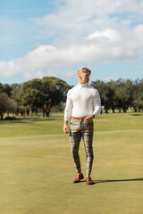 A blonde man walks on a green golf course in Miami, wearing a white turtleneck sweater and plaid pants.