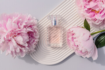 A bottle of women's perfume lies on a white podium in the form of an arch with delicate lilac peonies. top view. The concept of beauty and perfume.