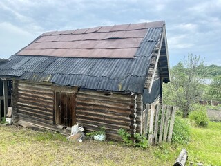 An old wooden cabin with a corrugated metal roof sits in a grassy clearing against a backdrop of greenery and an overcast sky. High quality photo