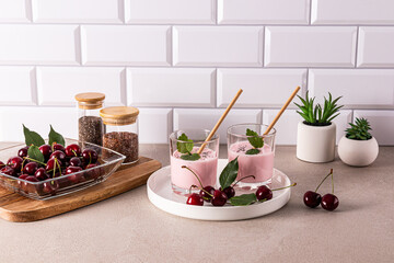 Delicious healthy cherry smoothie in two glass glasses with eco straw on a ceramic tray on a stone countertop with cooking ingredients.