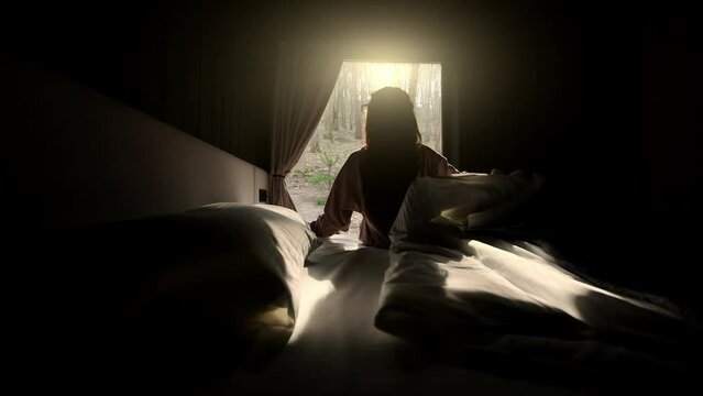Woman waking up in bed, looking out window at the morning sunlight in a forest setting.