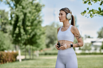 A young woman with vitiligo, wearing active wear and headphones, walks through a park during her workout.