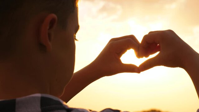 A close-up silhouette of a boy seen from behind, holding his hands in the shape of a heart. boy hand in sun rays hands at sunset, creating an atmosphere of childhood dreams and imagination.