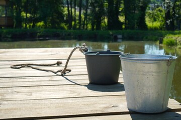 Naklejka premium buckets of metal and plastic on a wooden pier against the background of a lake in summer where carp are grown on a family farm