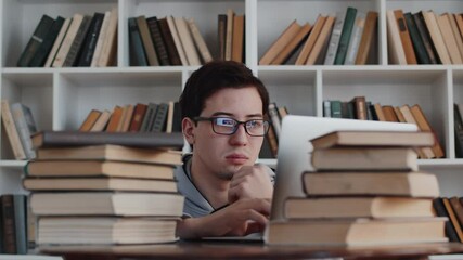 Young asian man in glasses doing his education, working on the laptop at home and getting tired, rubbing his eyes surrounded by stacks of books