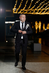 A man in a suit is walking down a street at night. He is wearing a tie and a boutonniere