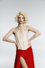 A drag queen with blonde curls and bold red lipstick poses in a white corset and a red, fuzzy skirt.