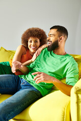 An African American man and woman, dressed vibrantly, share a moment of friendship while sitting on a yellow couch against a grey backdrop.