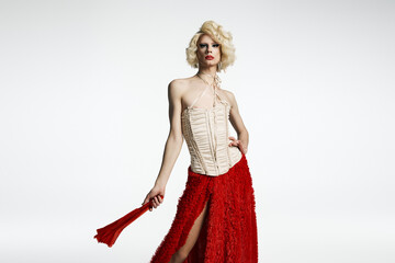 A drag queen in a blonde wig poses in a red skirt and white corset against a grey backdrop.