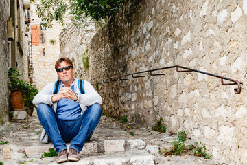 A traveler in sunglasses and a backpack relaxes in a peaceful alley with rustic architecture. Saint...