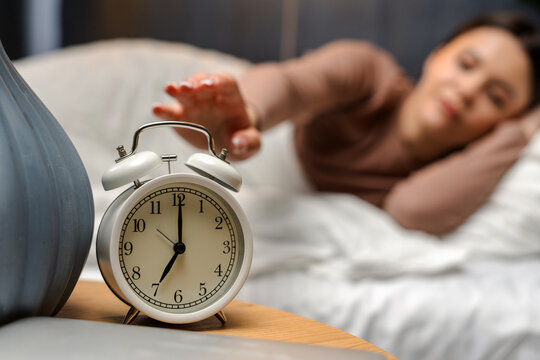 Young female turning off alarm clock on nightstand at 6am