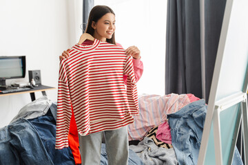 Young girl, teenager choosing clothes holding red striped shirt in messy room