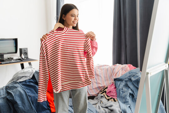 Young girl, teenager choosing clothes holding red striped shirt in messy room