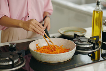 Close-up of young woman using sticks to fry carrot on pan on stove