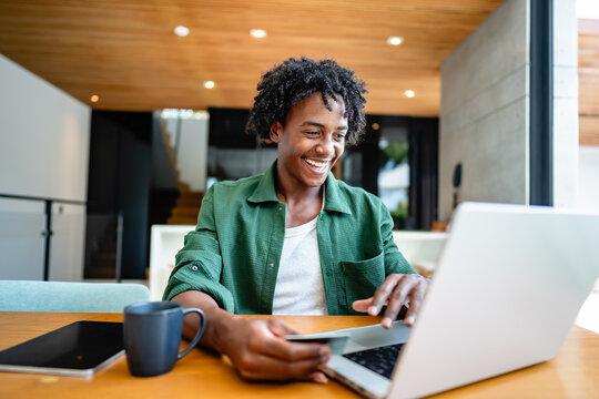 A young man of African descent sits by a wooden table in a modern, sunlit living room, smiling as he uses a laptop to make an online purchase and pays by credit card.