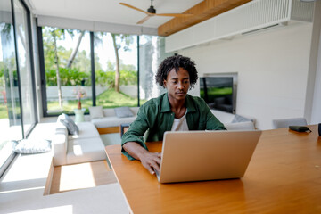 A young man of African descent sitting in a modern living room, looking at the laptop. His...