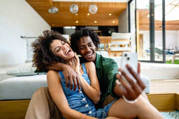 A young couple of African descent is sitting in a modern living room, both smiling and enjoying...